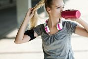 Eine junge Frau in Fitnesskleidung trinkt aus einer pinkigen Wasserflasche und hält ihren blonden Pferdeschwanz mit der anderen Hand. Sie trägt ein graues T-Shirt, pink-weisse Kopfhörer um ihren Hals und eine Armband für ihr Smartphone. Im Hintergrund sieht man verschwommen eine urbane Umgebung.  Eine junge Frau in Fitnesskleidung trinkt aus einer pinkigen Wasserflasche und hält ihren blonden Pferdeschwanz mit der anderen Hand. Sie trägt ein graues T-Shirt, pink-weisse Kopfhörer um ihren Hals und eine Armband für ihr Smartphone. Im Hintergrund sieht man verschwommen eine urbane Umgebung.